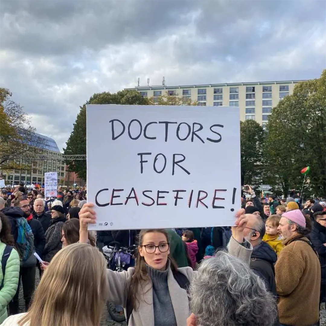 Frau hält bei einer Demonstration unter grauem Himmel ein handgeschriebenes Protestschild hoch, dahinter eine dichte Menschenmenge – ärztliche Stimme für Frieden